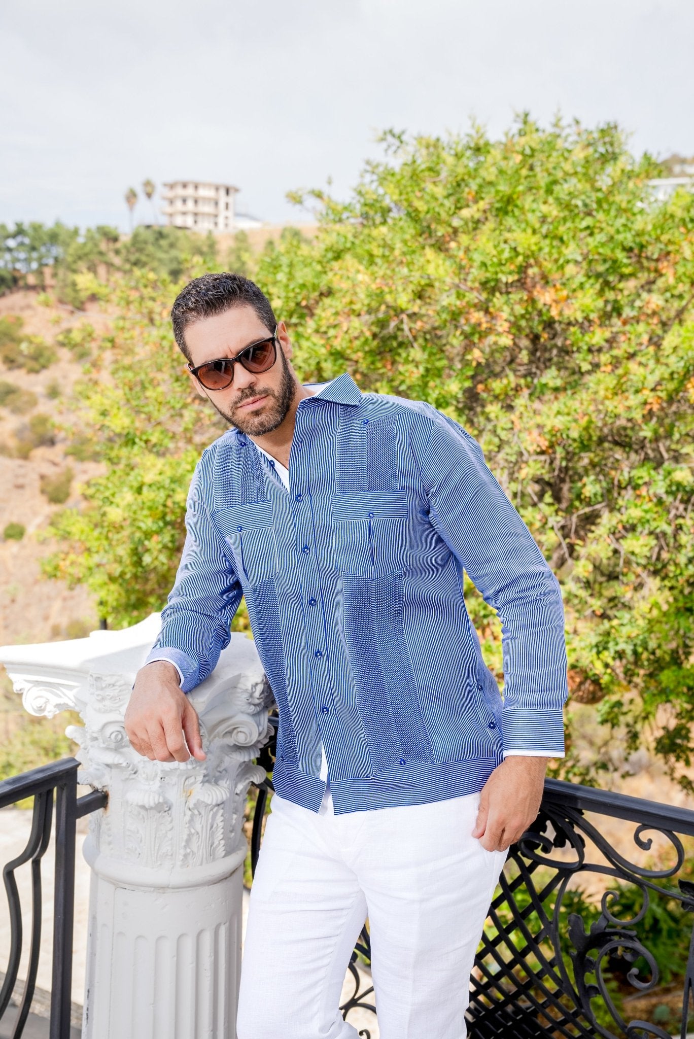 A man leans against a pillar wearing the blue and white, long-sleeved 100% Linen Contrast Panel Guayabera Shirt with white pants.