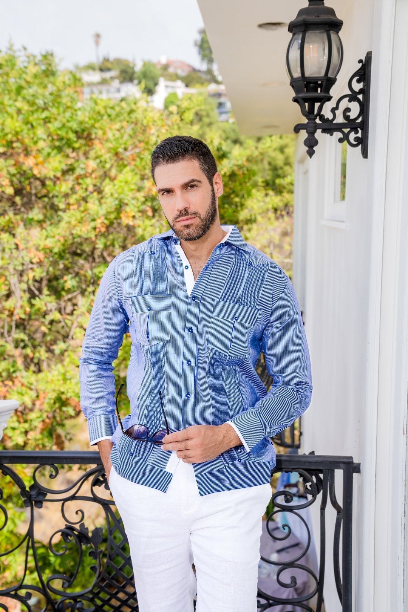 A man wears a blue striped 100% Linen Contrast Panel Guayabera Shirt with long sleeves and white linen pants.