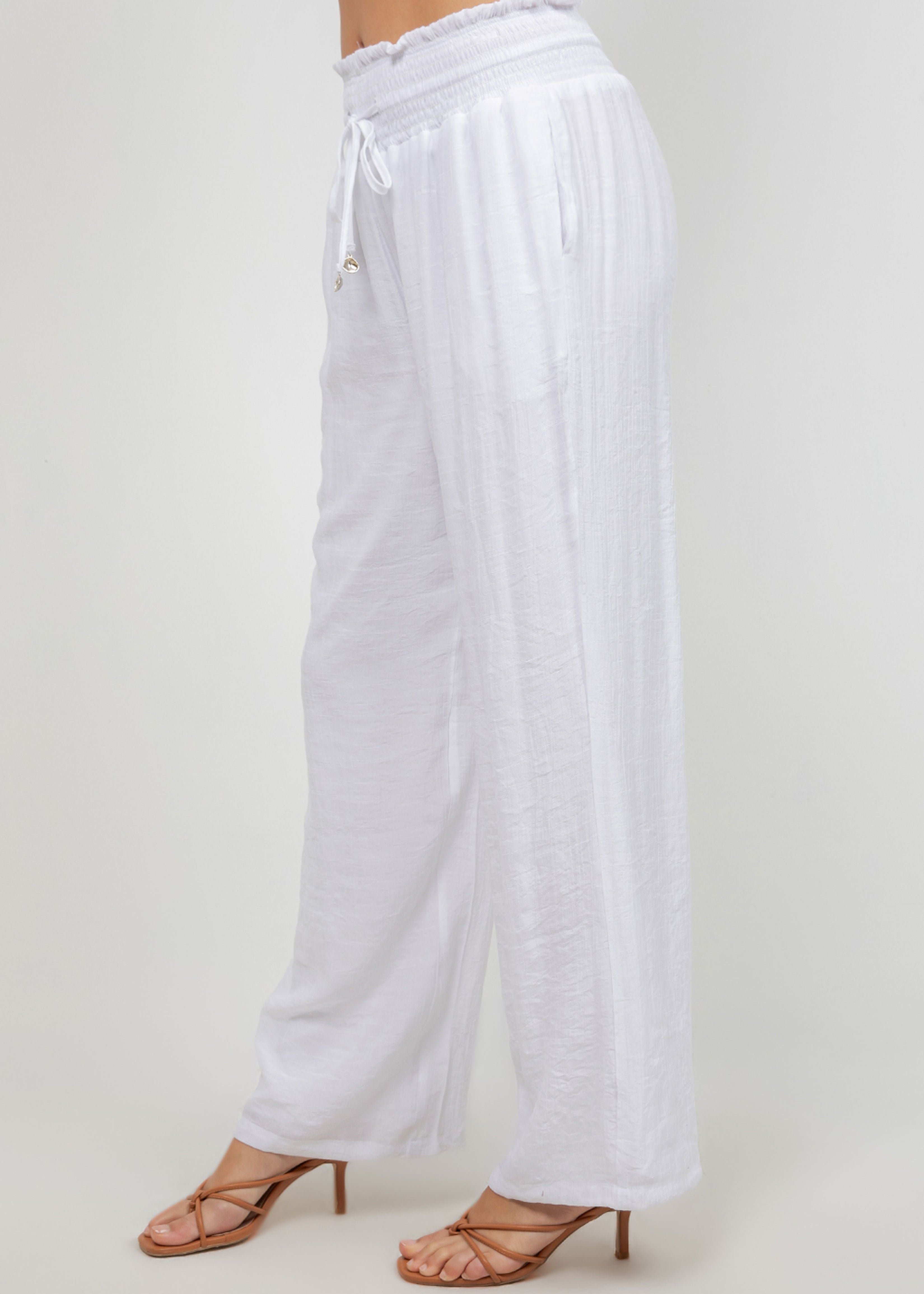 Side view of model wearing white Ladies Smocked Waist Drawstring Palazzo Pants and strappy brown heels.