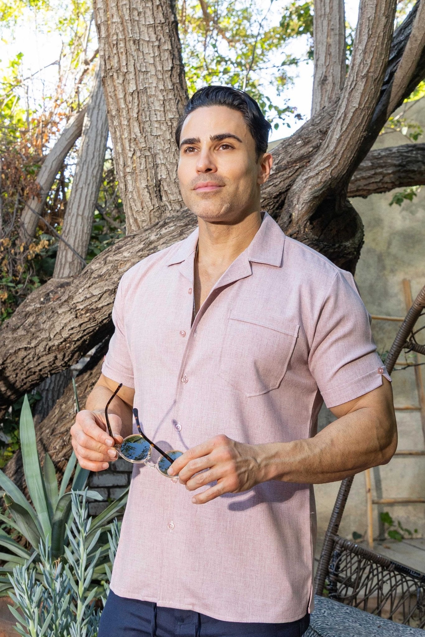 A man wears a pink Menâ€™s Retro Cuban Shirt with pleating and one pocket, holding sunglasses outside.
