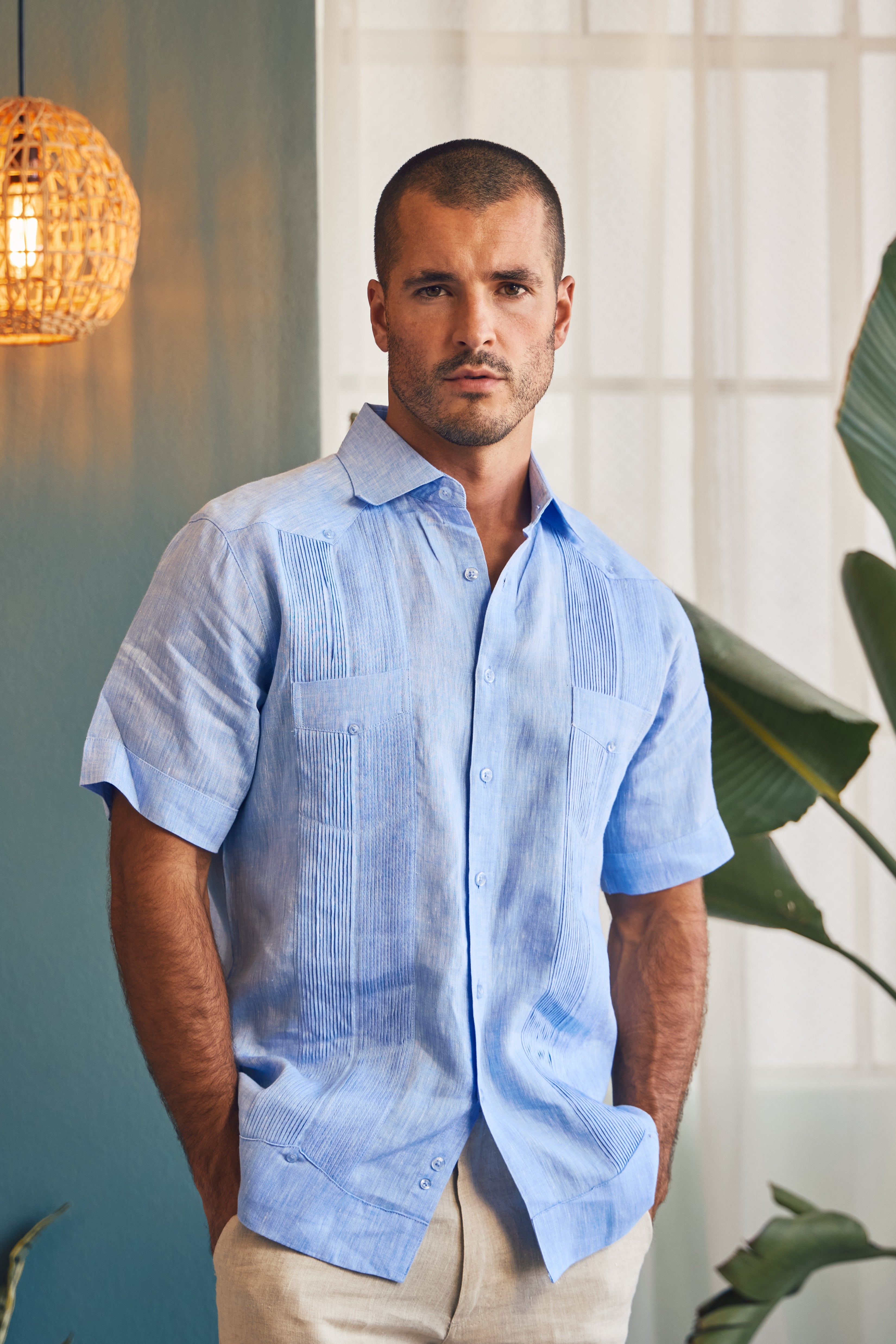 Man wearing a light blue shirt standing indoors with a plant and lamp in the background