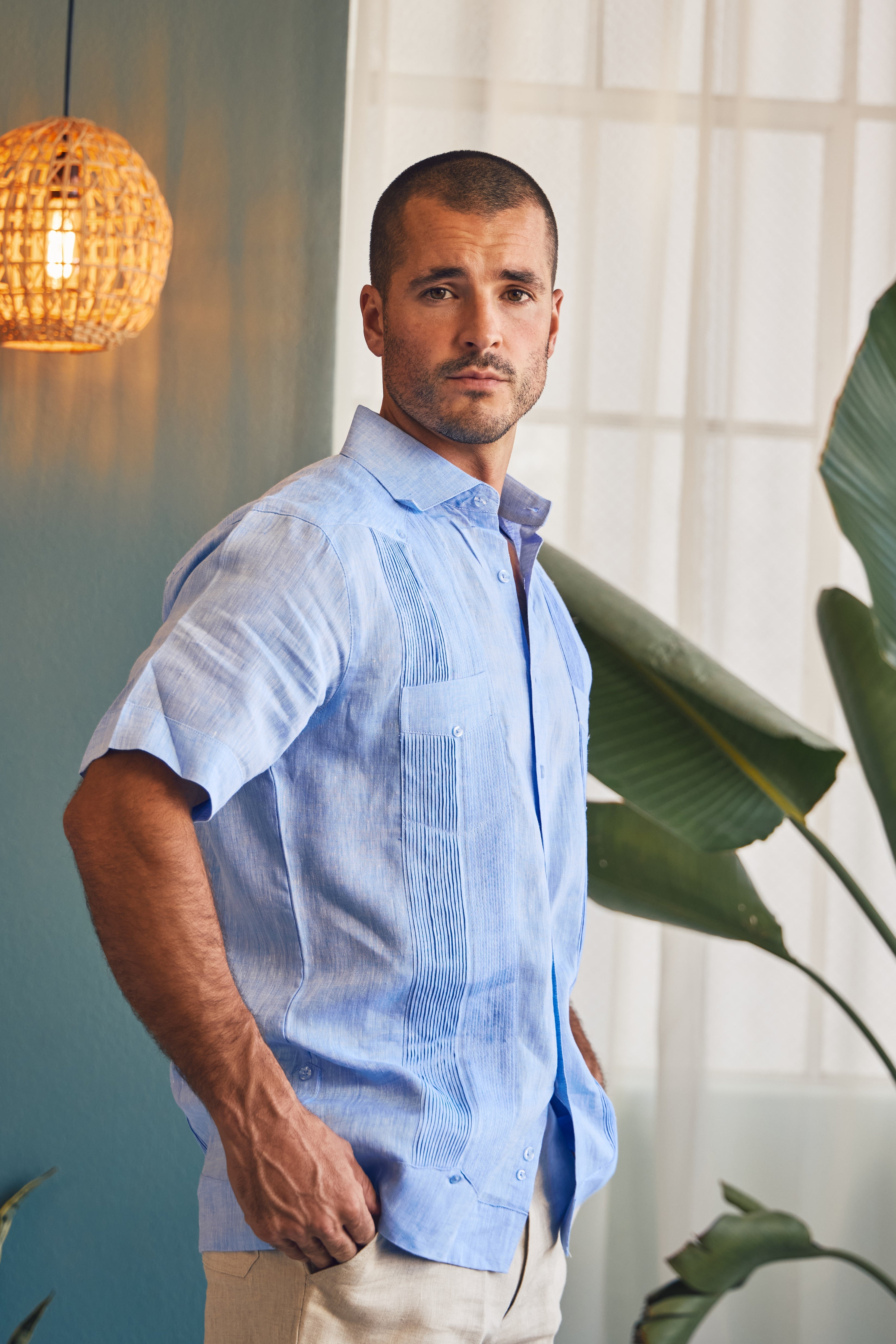 Man wearing a light blue shirt standing indoors with plants and a lamp in the background