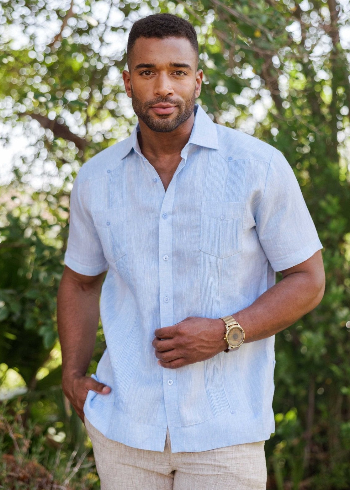 A man models a light blue Yarn Dye 100% Linen Two Pocket Guayabera Short Sleeve shirt, with visible vertical pleats and two front pockets.