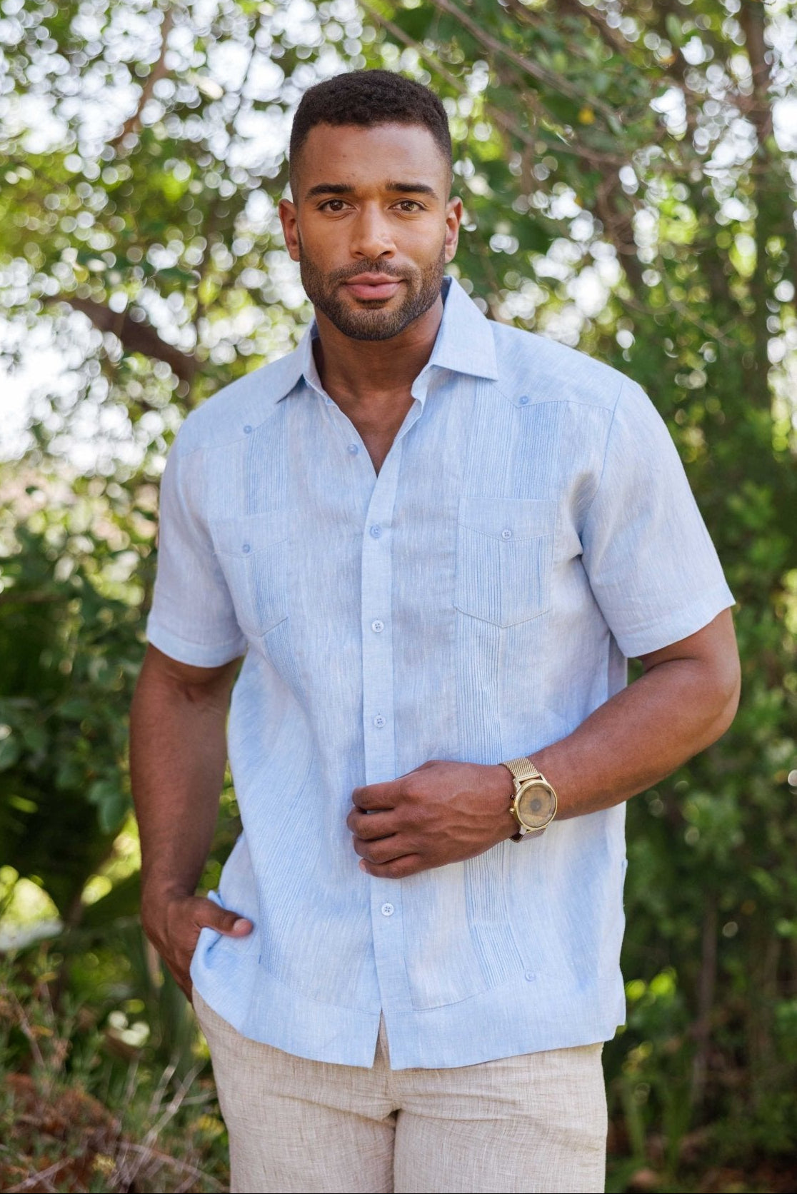 A man models a light blue Yarn Dye 100% Linen Two Pocket Guayabera Short Sleeve shirt, with visible vertical pleats and two front pockets.