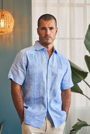Man wearing a light blue shirt standing indoors with a plant and lamp in the background