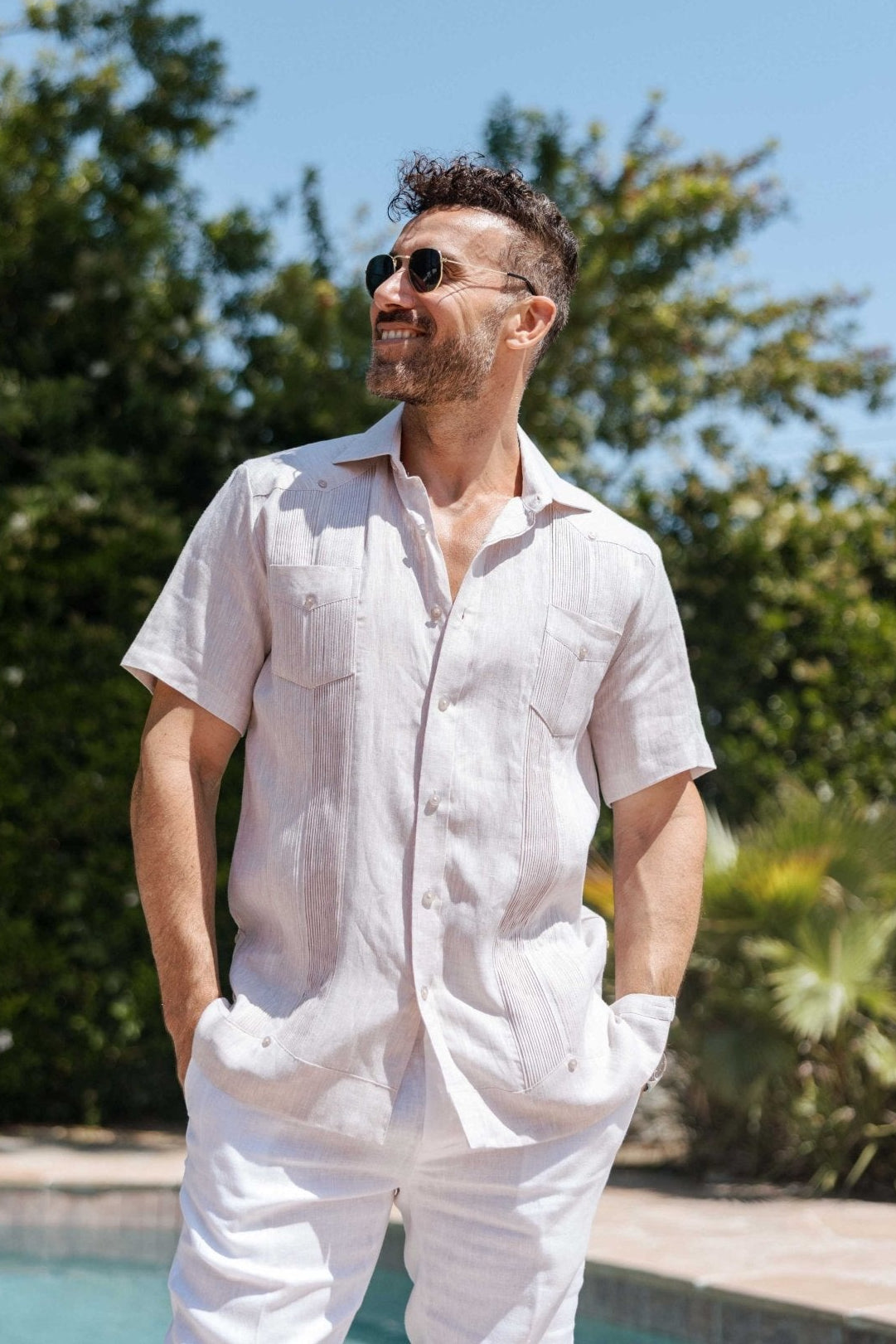 A man wears a tan Yarn Dye 100% Linen Two Pocket Guayabera Short Sleeve shirt with two chest pockets and white pants.