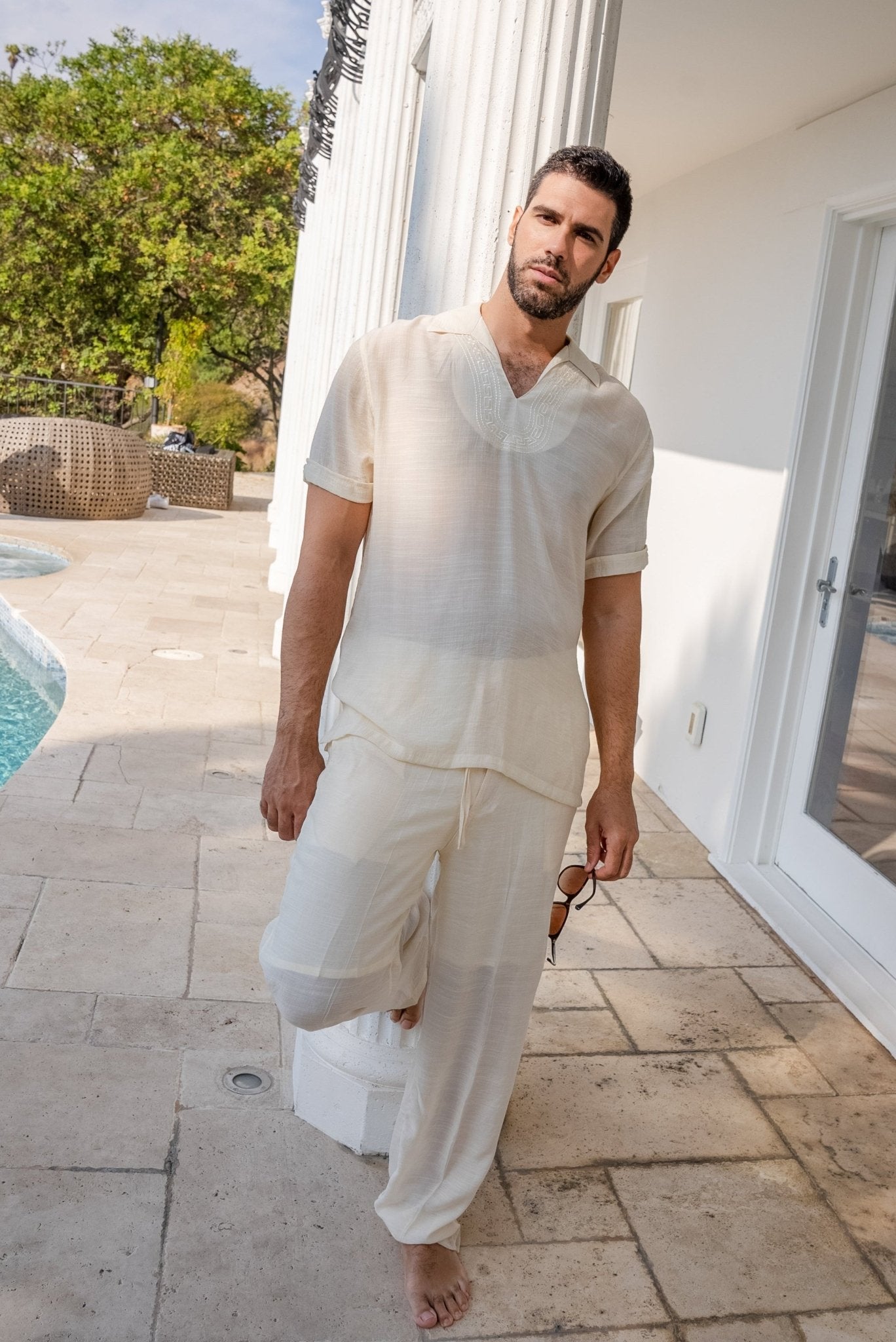 A man wearing a matching off-white short-sleeve V-neck t-shirt and wide-leg trousers stands barefoot on a stone patio next to a swimming pool and white building exterior.