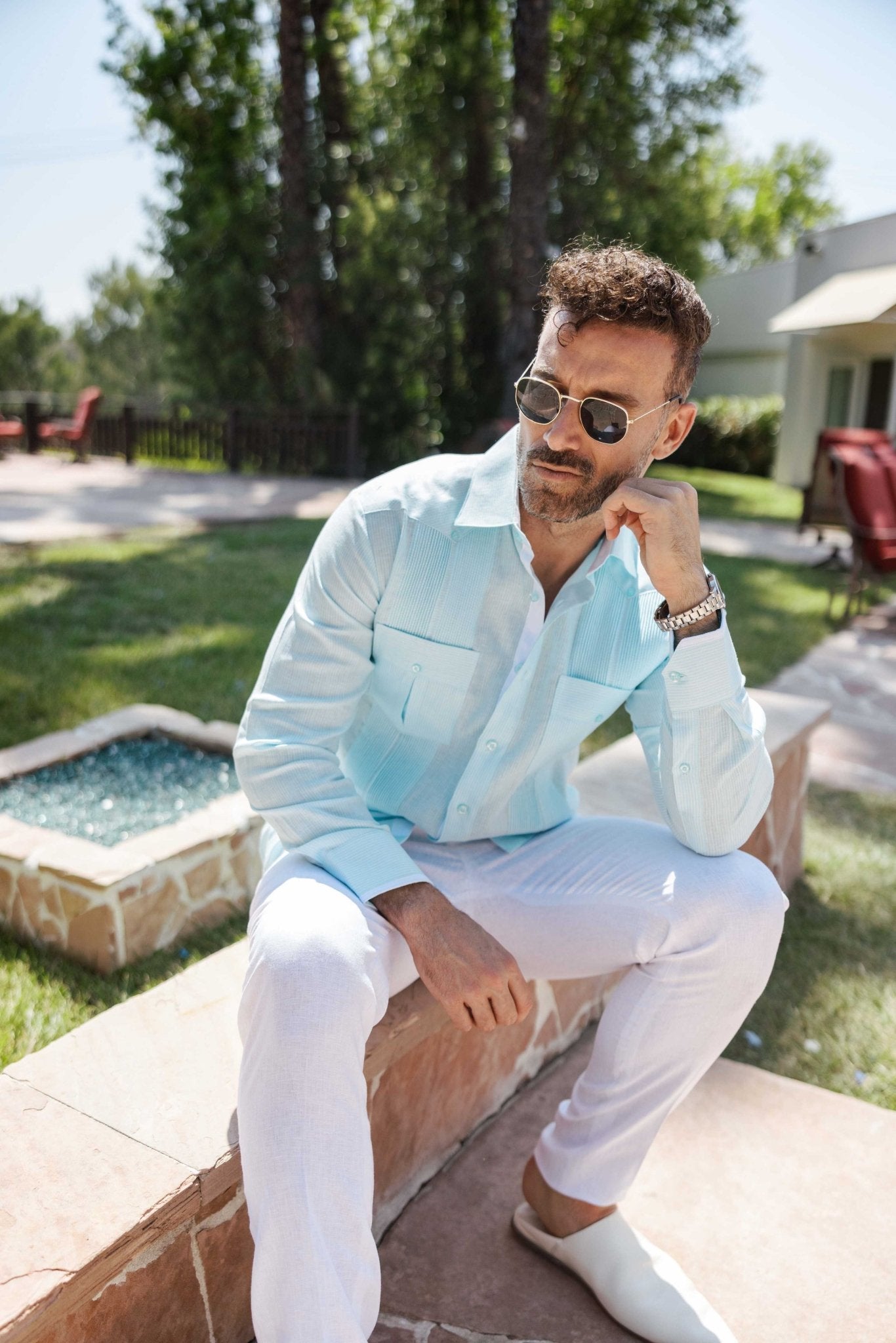 A man wearing a light blue 100% Linen Contrast Panel Guayabera Shirt with white linen pants poses outside.