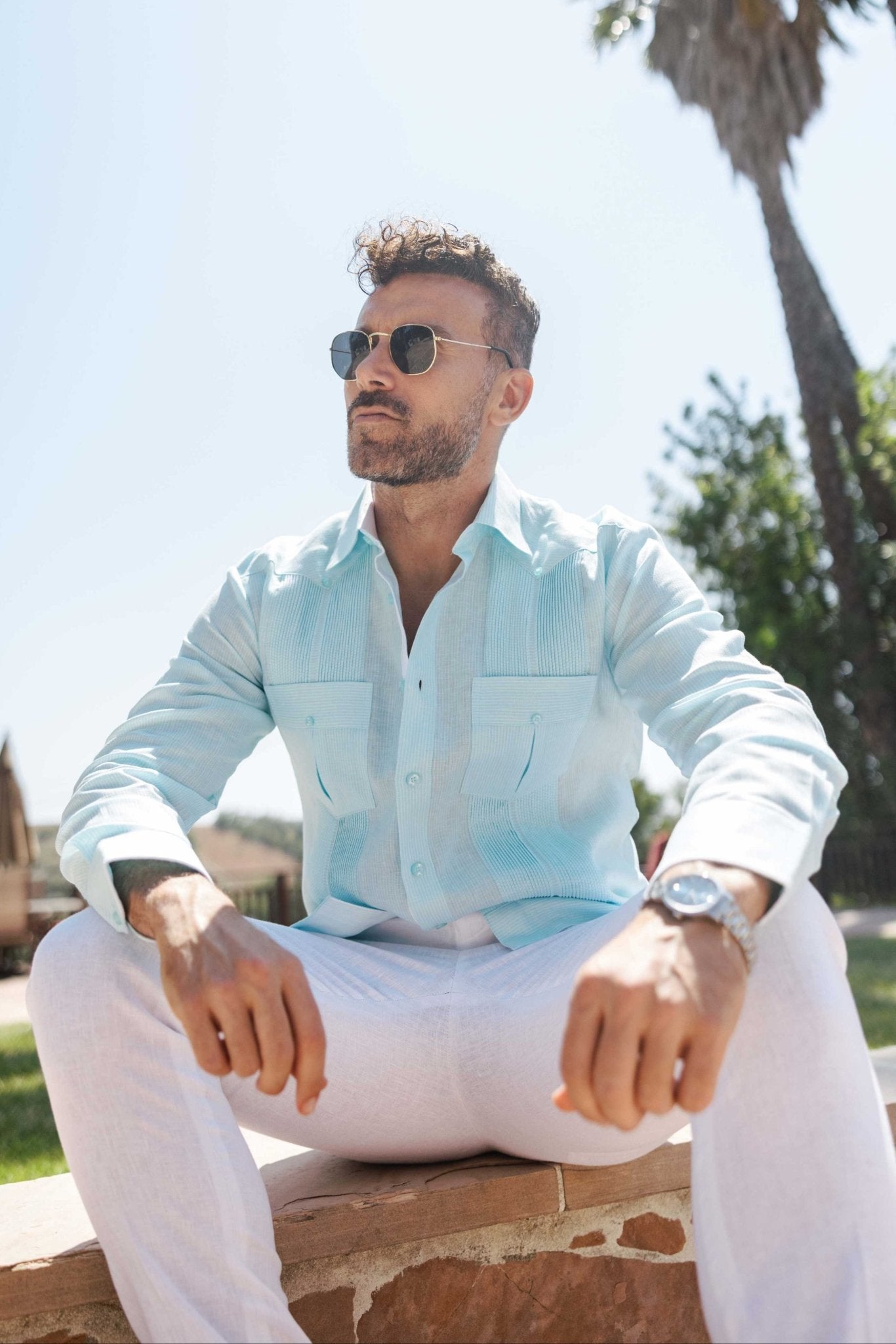 A man in sunglasses sits wearing the 100% Linen Contrast Panel Guayabera Shirt in aqua with long sleeves and white linen pants.