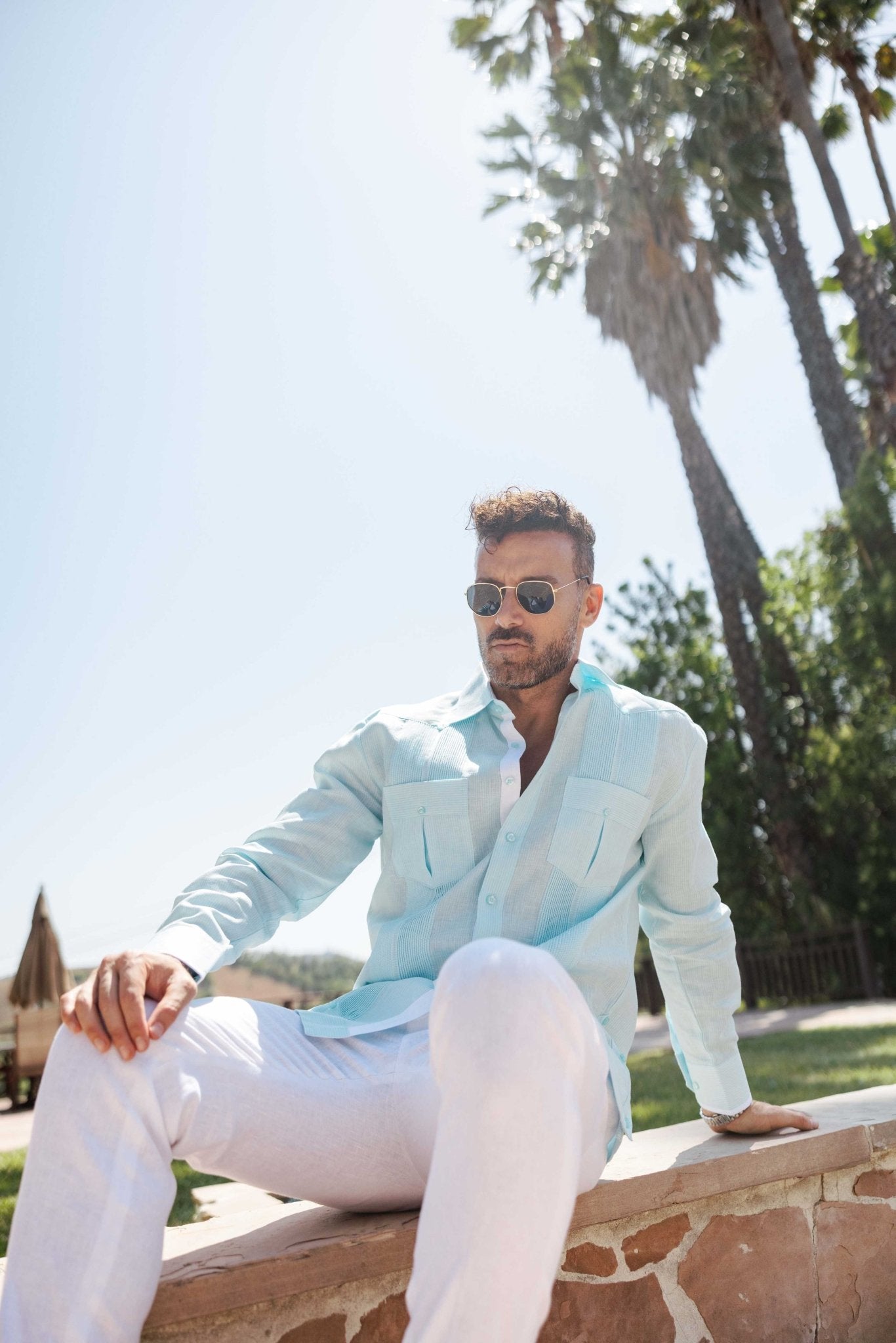 A man in sunglasses wears the light blue 100% Linen Contrast Panel Guayabera Shirt, seated, in a relaxed outdoor setting.