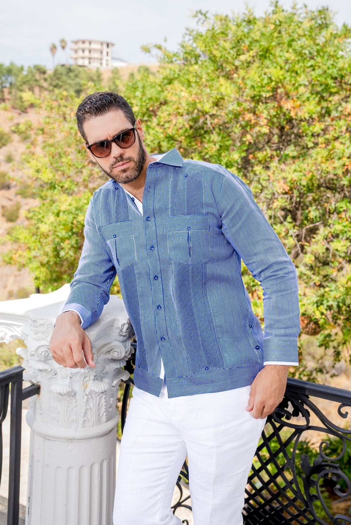 A man leans against a pillar wearing the blue and white, long-sleeved 100% Linen Contrast Panel Guayabera Shirt with white pants.
