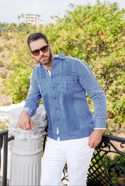 A man leans against a pillar wearing the blue and white, long-sleeved 100% Linen Contrast Panel Guayabera Shirt with white pants.