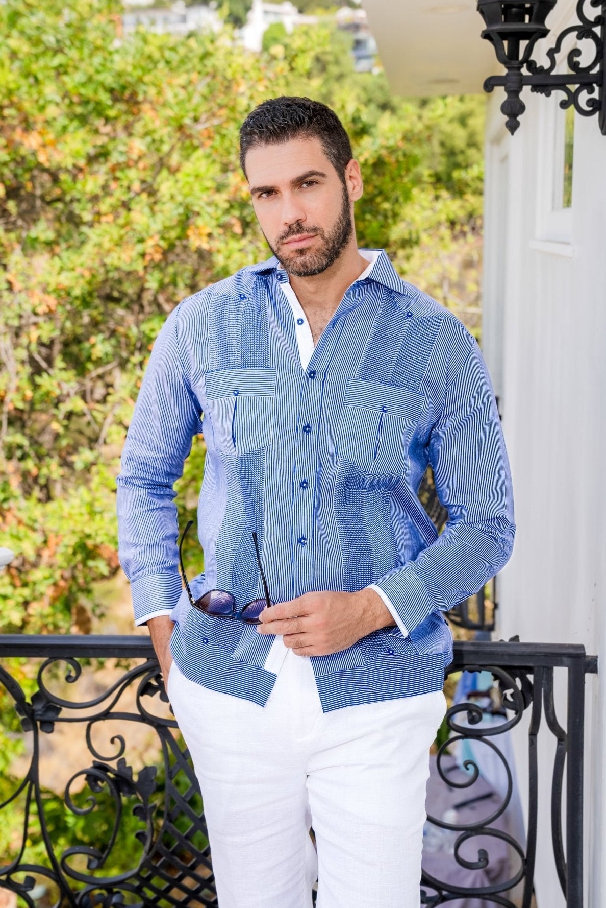 A man wears a blue striped 100% Linen Contrast Panel Guayabera Shirt with long sleeves and white linen pants.