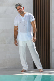 A man wearing a white short-sleeve linen tunic top with a lace-up neckline and matching white linen trousers, standing barefoot near a pool in front of a light stone wall and dark brown slatted door.