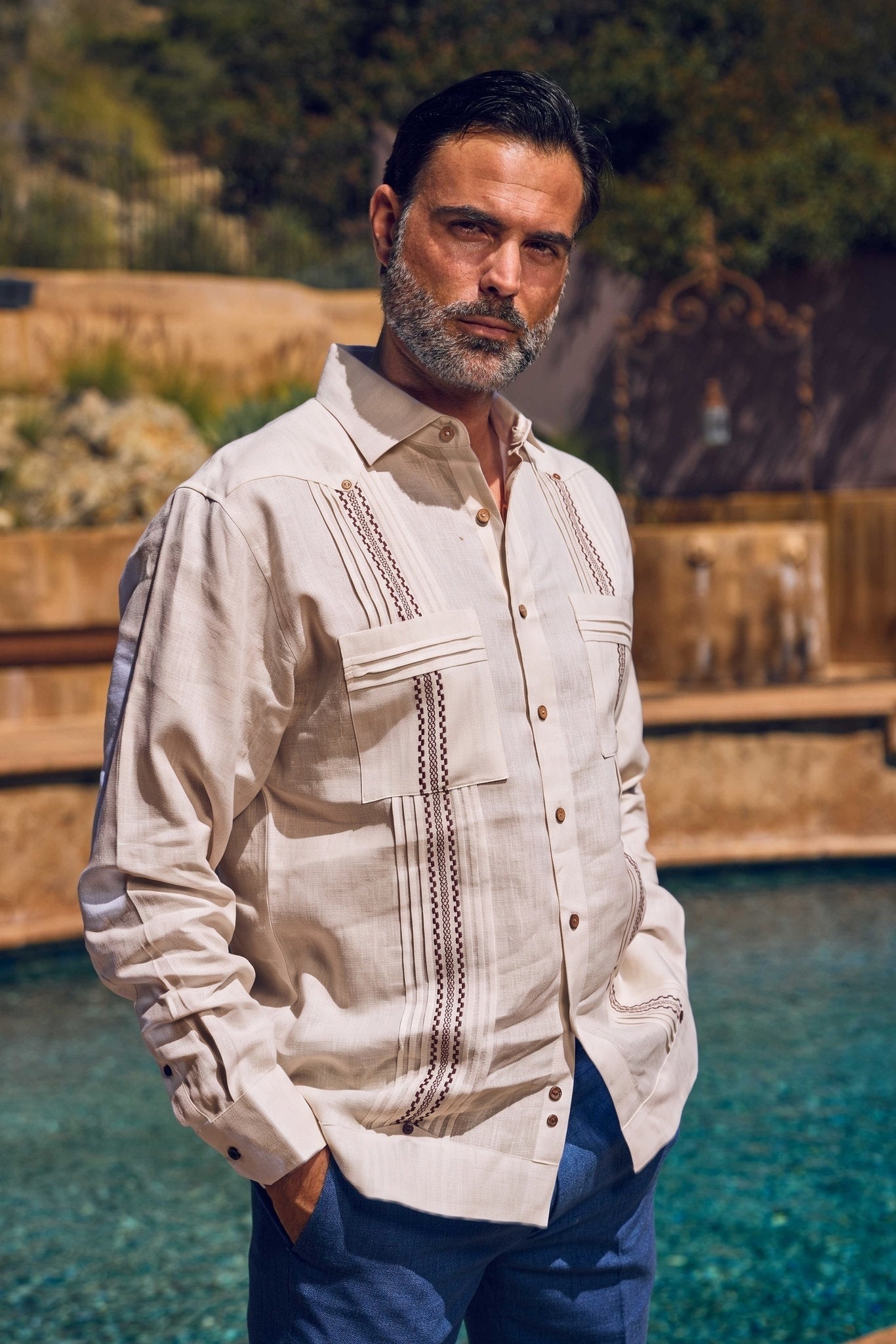 A man wearing a light beige Guayabera-style shirt with brown embroidered details and blue pants, standing outdoors near a pool with a blurred stone and green background.