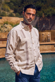 A man wearing a light beige Guayabera-style shirt with brown embroidered details and blue pants, standing outdoors near a pool with a blurred stone and green background.