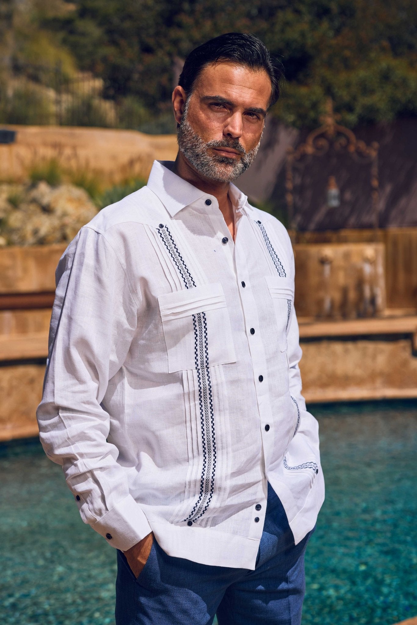Man models the white 100% Linen Embroidery Trimmed Guayabera Long Sleeve with blue embroidery detail; chest up shot.
