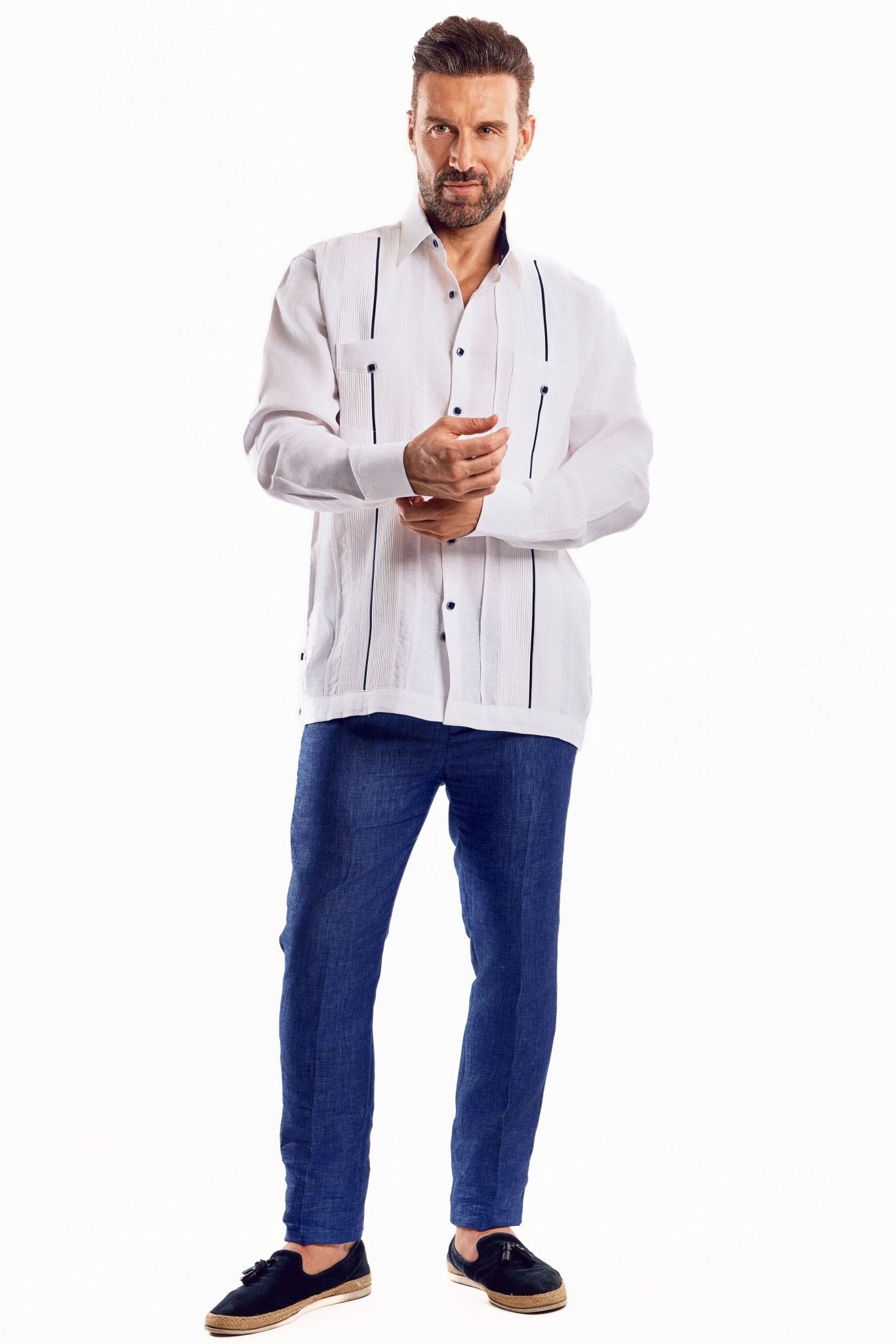A man wears a white linen long-sleeve Guayabera shirt with contrast trim, paired with blue pants.
