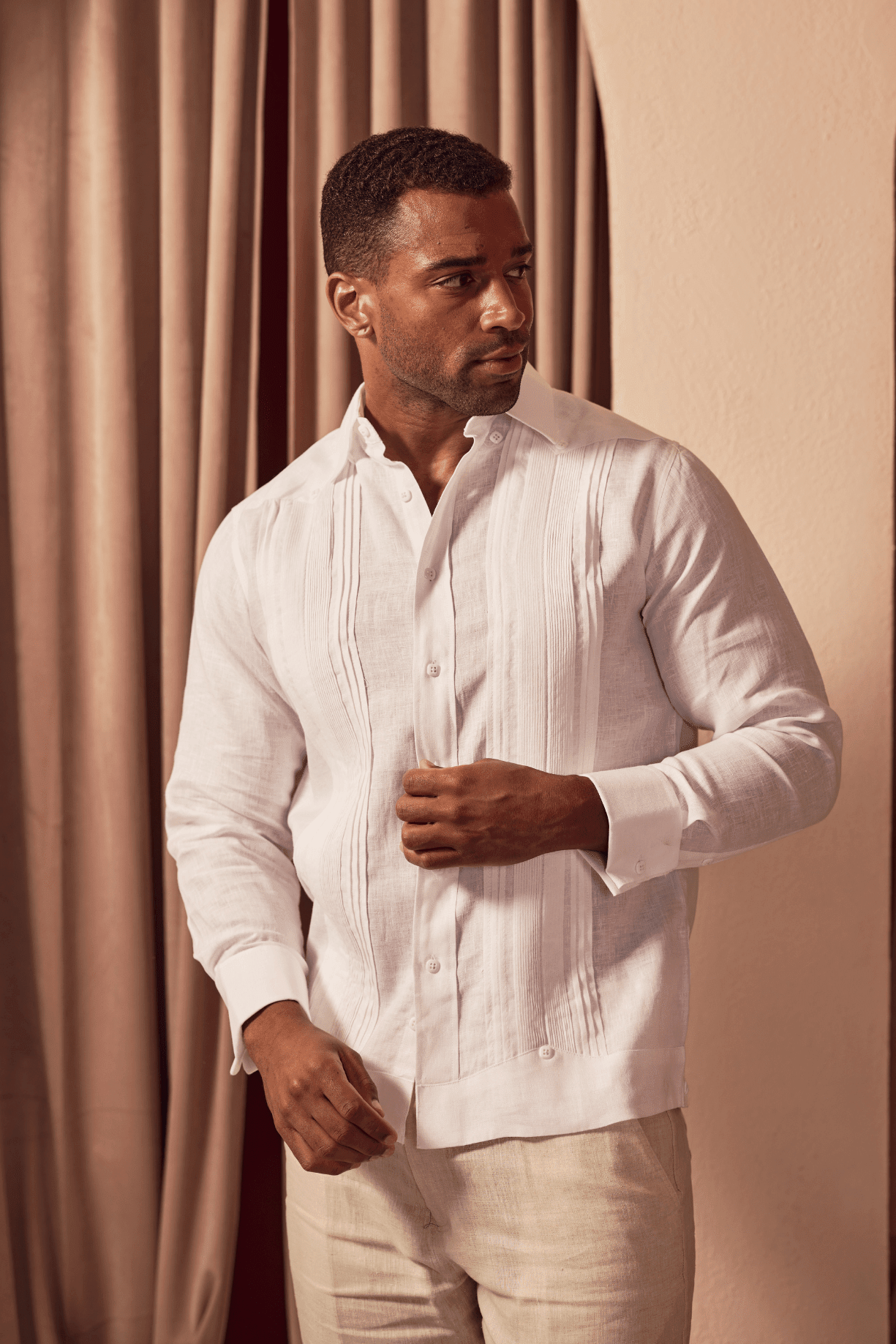 A man wearing a white guayabera-style button-up shirt and light beige trousers, standing in front of warm-toned curtains and an arched wall.