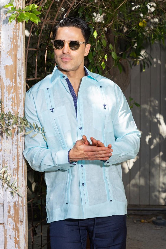 A man in a light blue Men's Long Sleeve Linen Guayabera Shirt leaning against a post, styled with sunglasses for a tropical event.