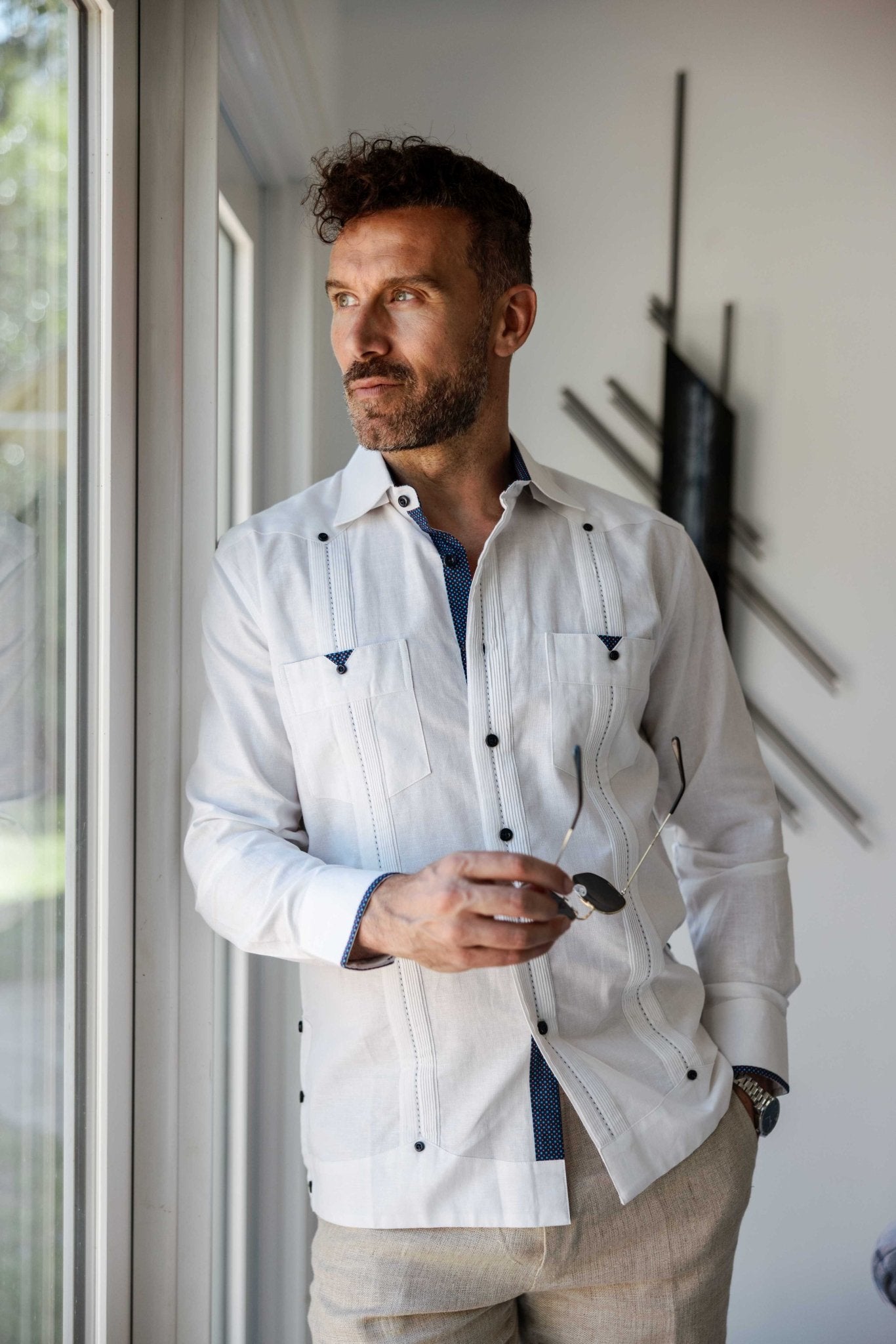 A man wearing a white long sleeve linen guayabera shirt with blue printed trim standing by a window, styled with khaki linen pants for a relaxed resort look.