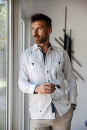 A man wearing a white long sleeve linen guayabera shirt with blue printed trim standing by a window, styled with khaki linen pants for a relaxed resort look.