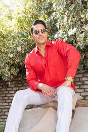 A man models the red, long-sleeve, multi-pleat Yarn Dyed Linen Guayabera, while sitting and wearing silver rimmed sunglasses.
