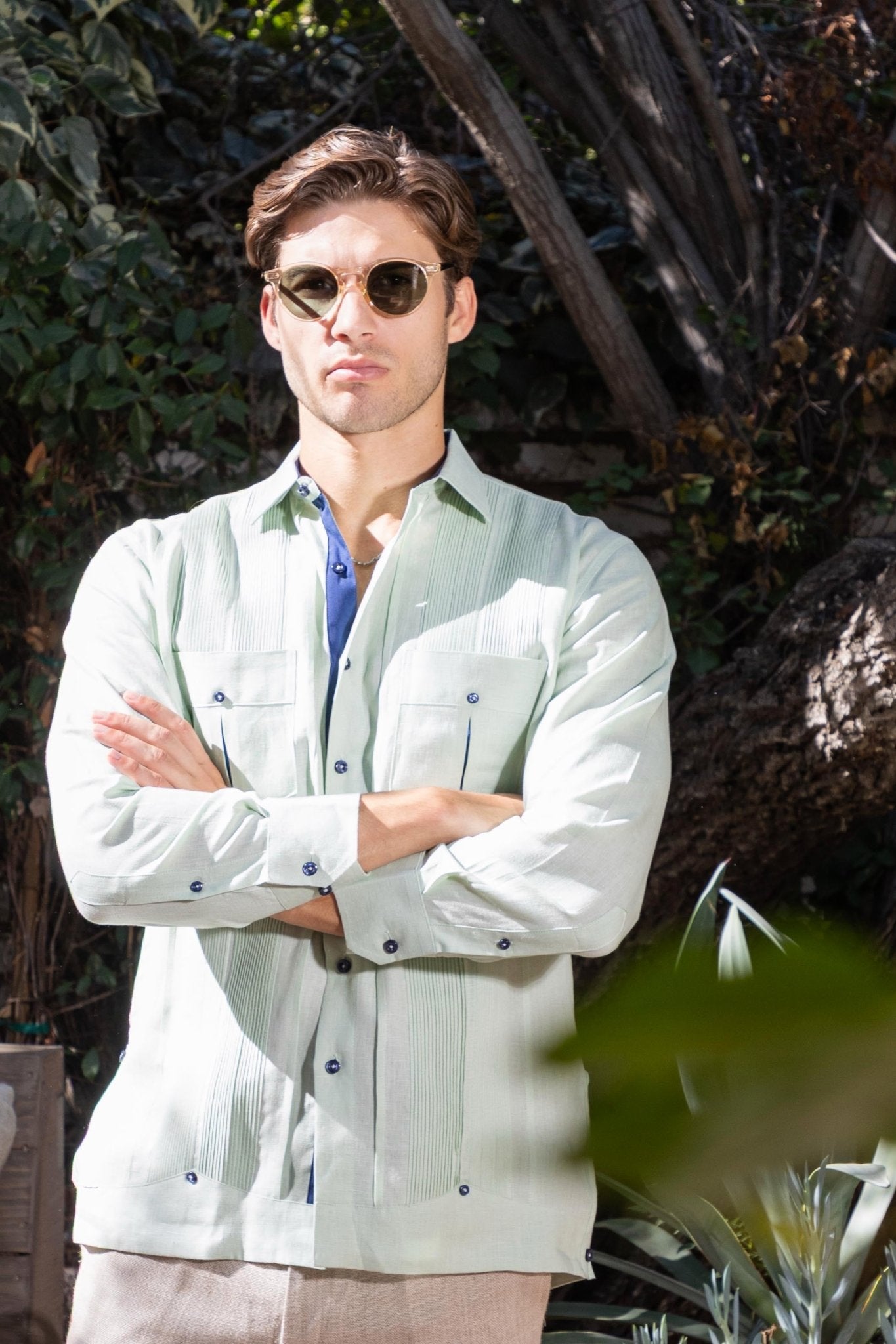 Men's long sleeve sage linen guayabera shirt with navy buttons and contrast detail, model arms-crossed wearing sunglasses in outdoor tropical garden setting