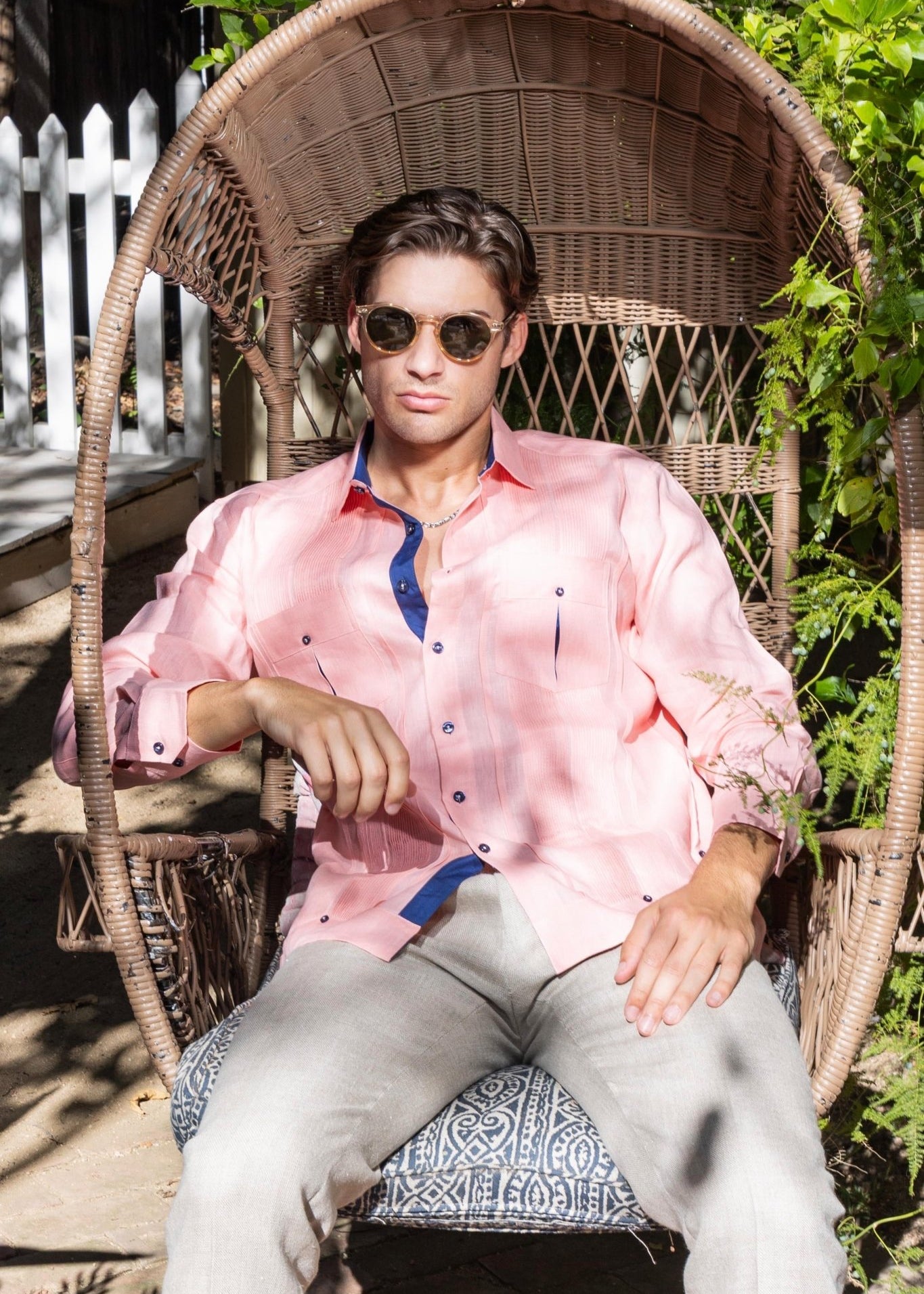 Men's linen guayabera shirt in salmon pink with navy trim, model wearing sunglasses seated in wicker chair showing chest pocket and pleated front detail in garden