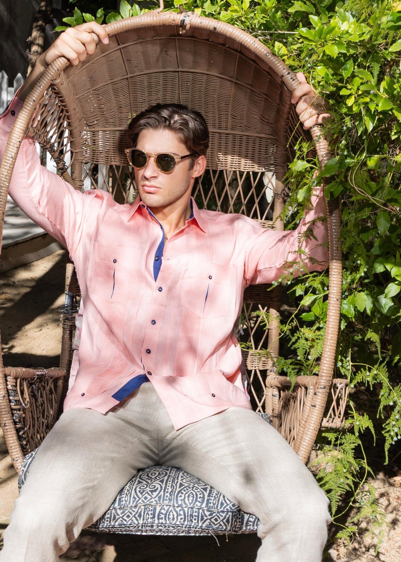 Men's premium linen guayabera shirt in salmon pink with navy buttons, model relaxing arms-up in rattan peacock chair surrounded by tropical greenery