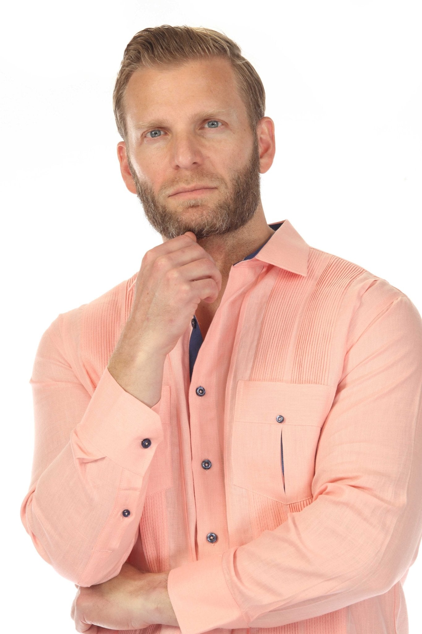 Men's salmon guayabera linen shirt portrait, model posing with hand on chin showing navy contrast collar and pleated chest panels in contemplative white background shot