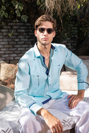 A man sits wearing a light blue, long-sleeved 100% Linen Contrast Panel Guayabera Shirt with dark blue accents.