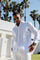 A man wears a white 100% Linen Contrast Panel Guayabera Shirt in front of palm trees.