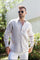A man wearing a beige long sleeve linen guayabera with white trousers standing outdoors.