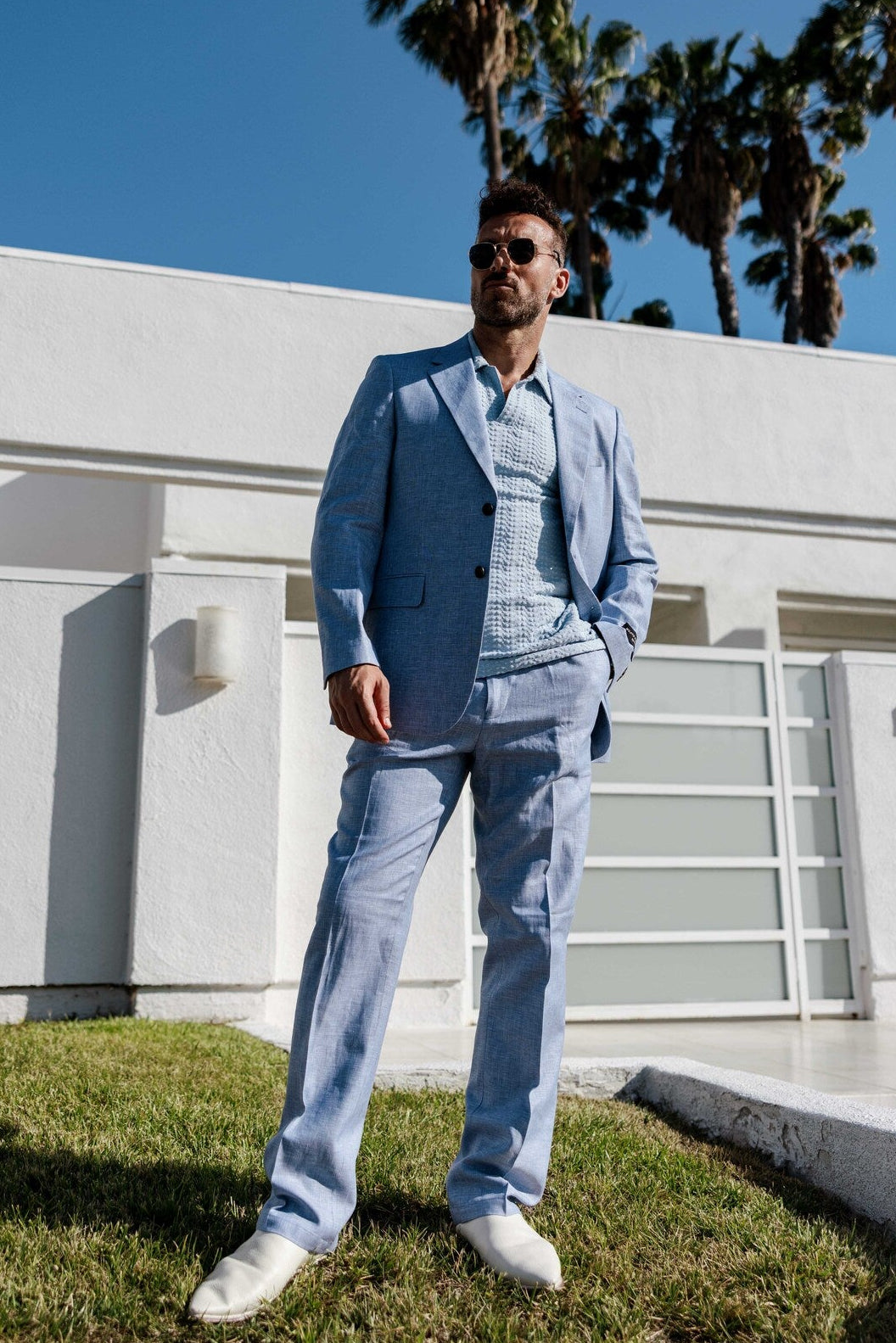 Man in a light blue suit standing in front of a modern white building with palm trees in the background.