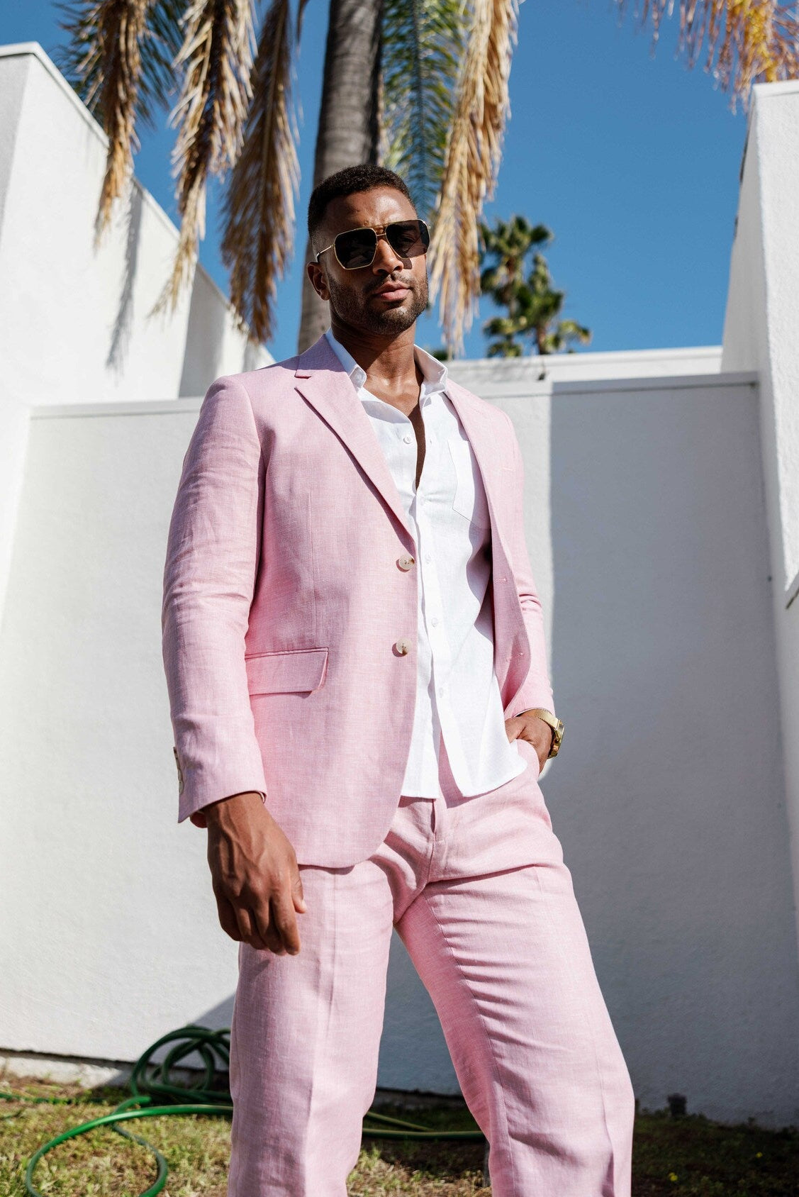 Man in a pink suit standing outdoors with palm trees and a white wall in the background