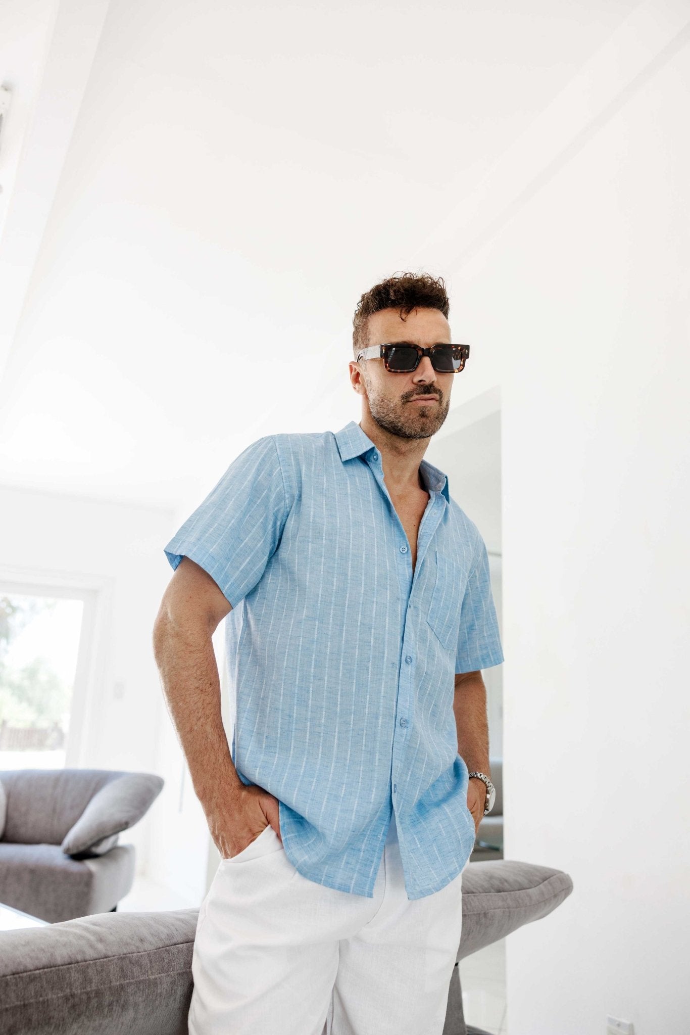 A man wears sunglasses with a light blue Menâ€™s Pinstripe Resort Shirt and white pants, posing with hands in his pockets.