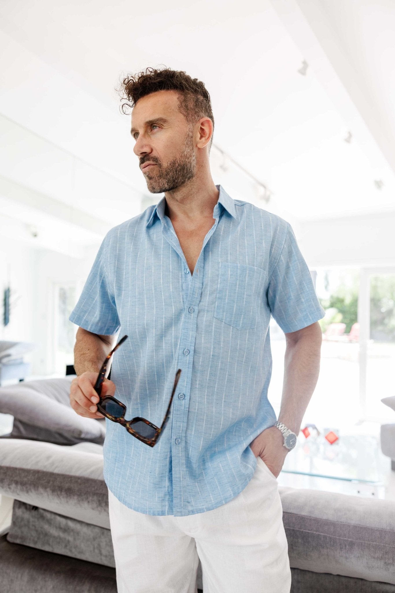 A man wears the Men’s Pinstripe Resort Shirt in light blue with white pinstripes, holding sunglasses.