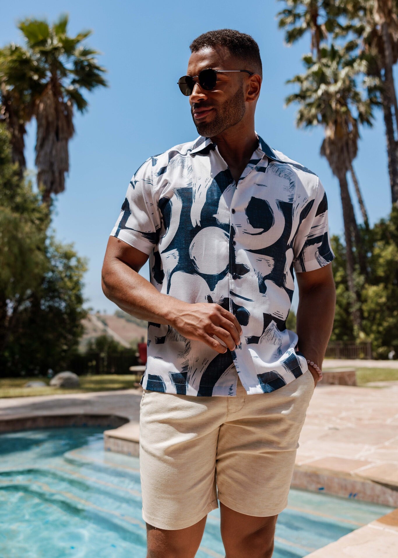 Man wearing a blue and white tropical print Men’s Resort Shirt and tan shorts stands by a pool in a medium shot.