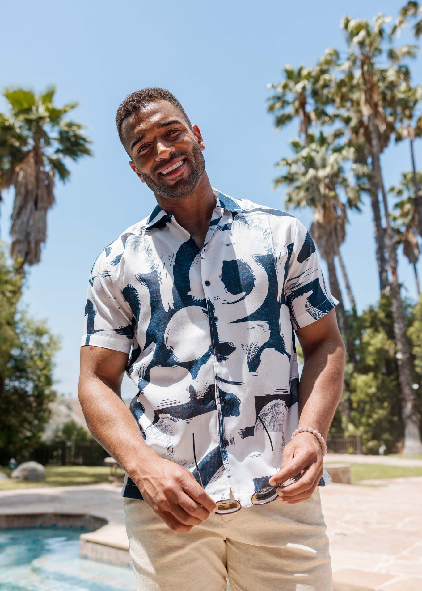 Smiling man holds sunglasses and wears the Men’s Tropical Print Resort Shirt with a blue abstract pattern in front of palm trees.
