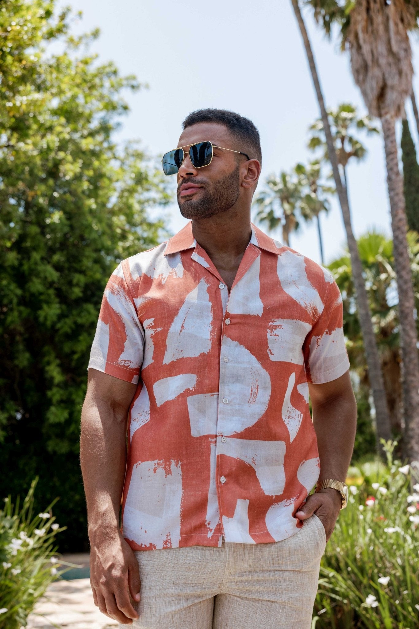 A man wears a tropical print resort shirt button-up in coral and white, looking to his left with palm trees in the background.