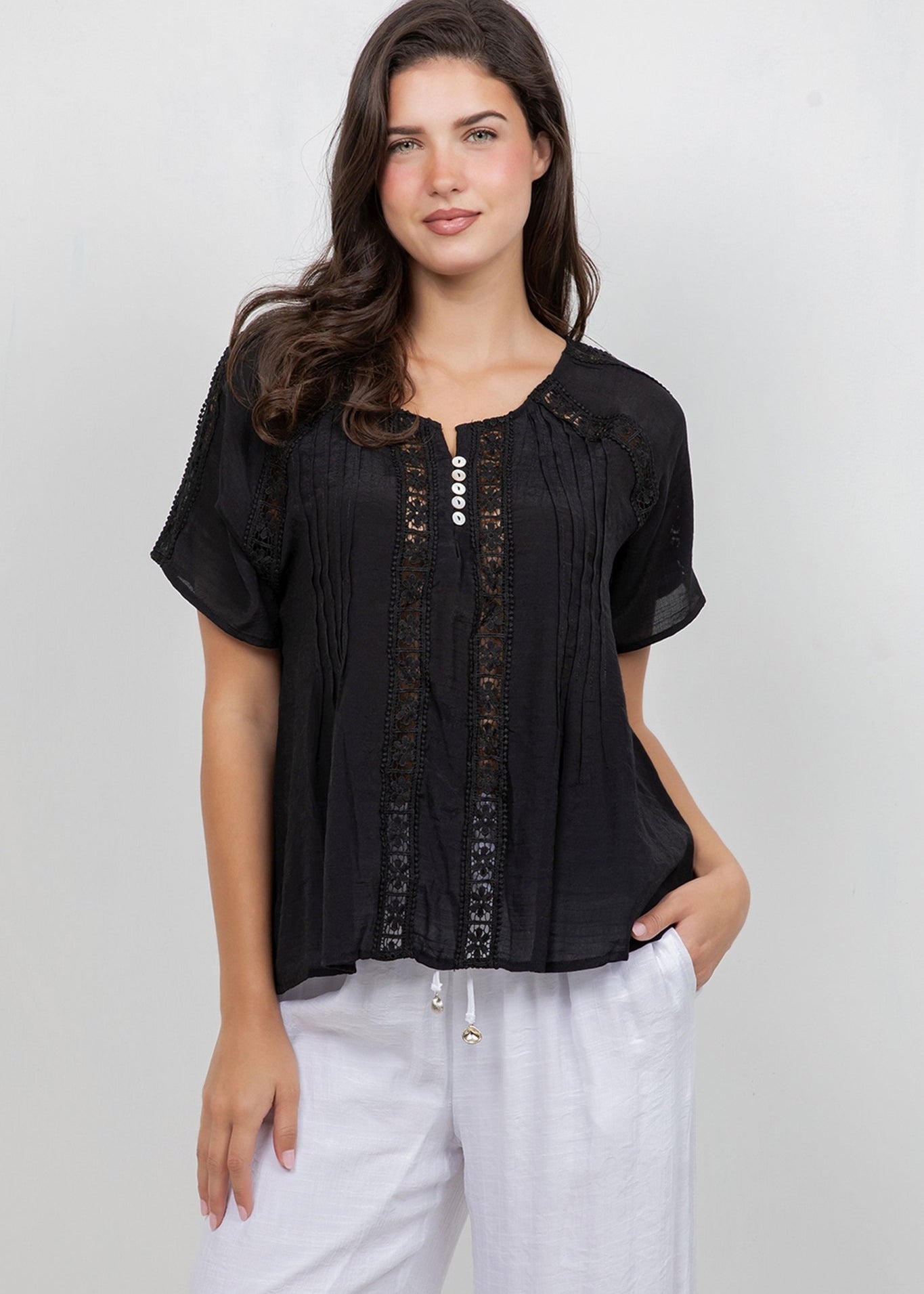 A woman models the Ladies Boho Pleated Lace Panel Blouse in black, which features white buttons and is styled with white pants.