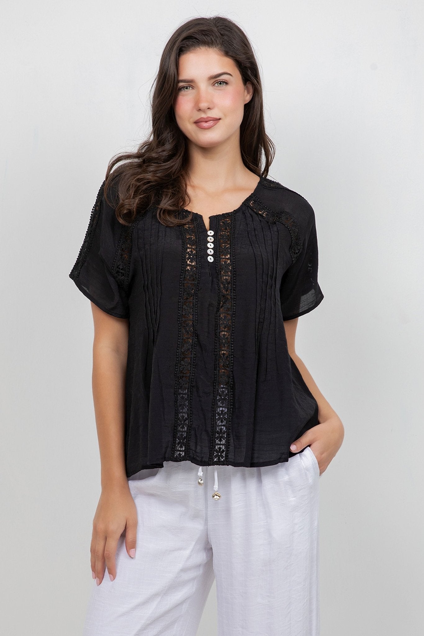 A woman models the Ladies Boho Pleated Lace Panel Blouse in black, which features white buttons and is styled with white pants.