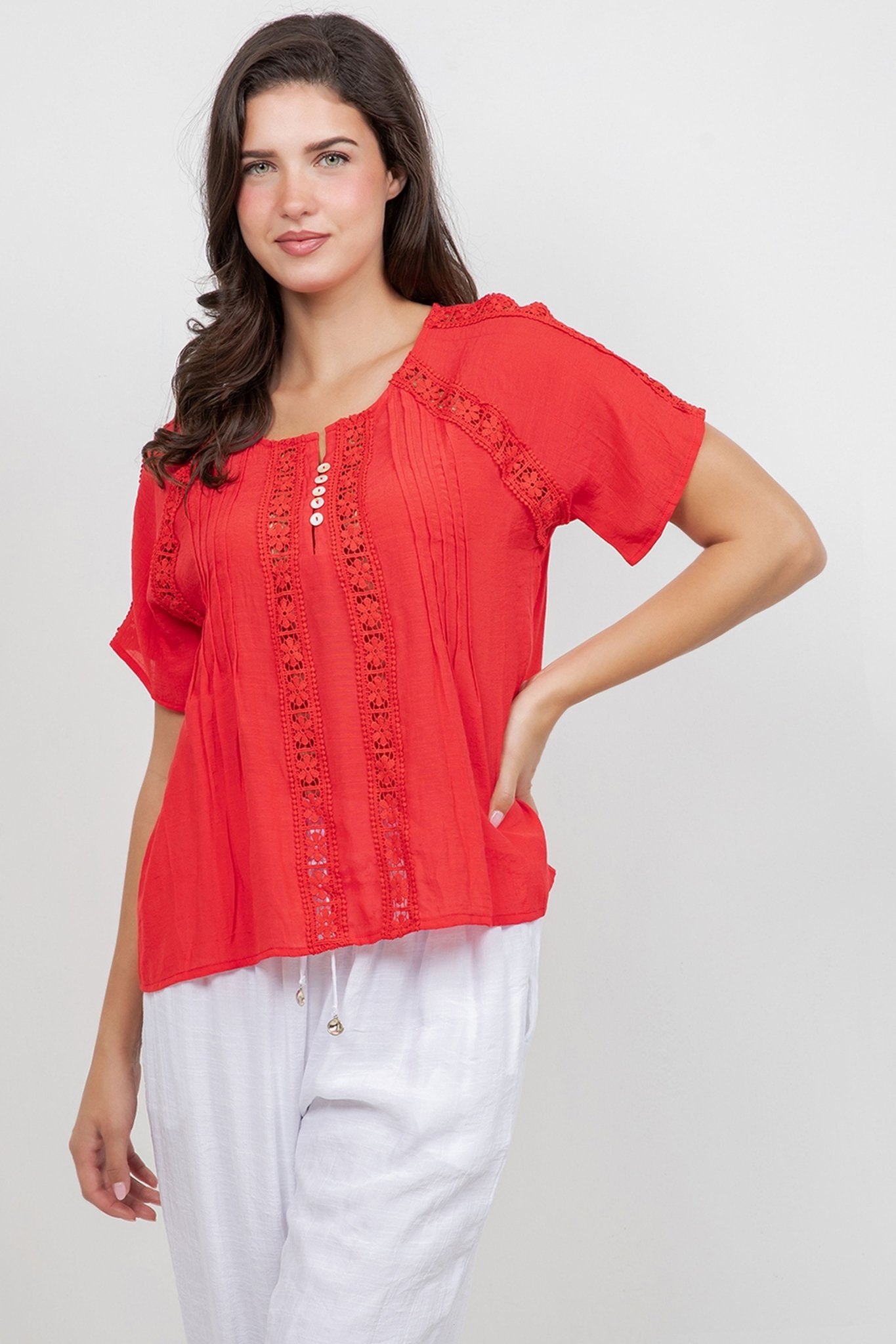 Front view of a model wearing a red Ladies Boho Pleated Lace Panel Blouse with white bottoms.