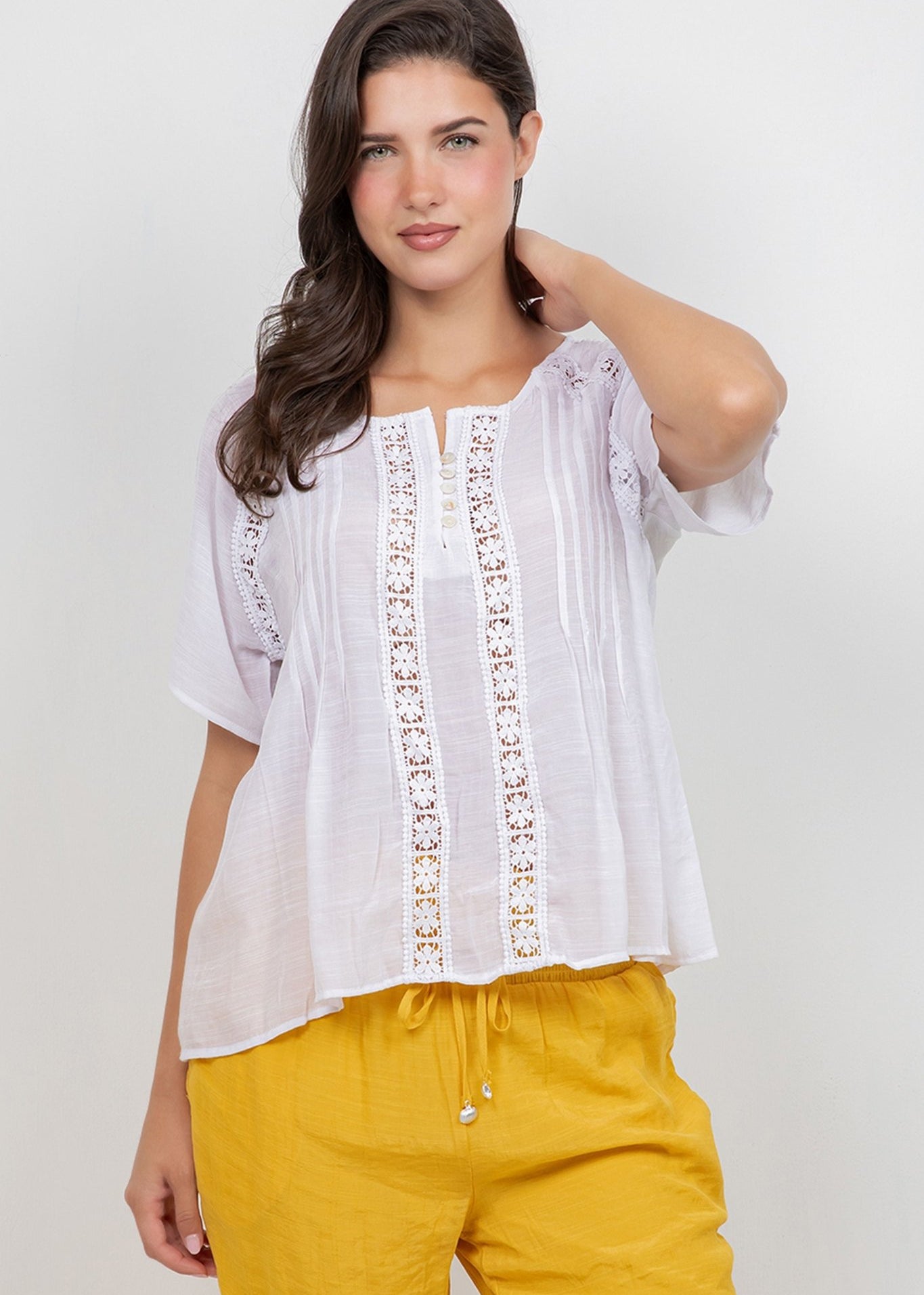 Woman wearing the white Ladies Boho Pleated Lace Panel Blouse, featuring lace panels and short sleeves, with mustard-colored pants.