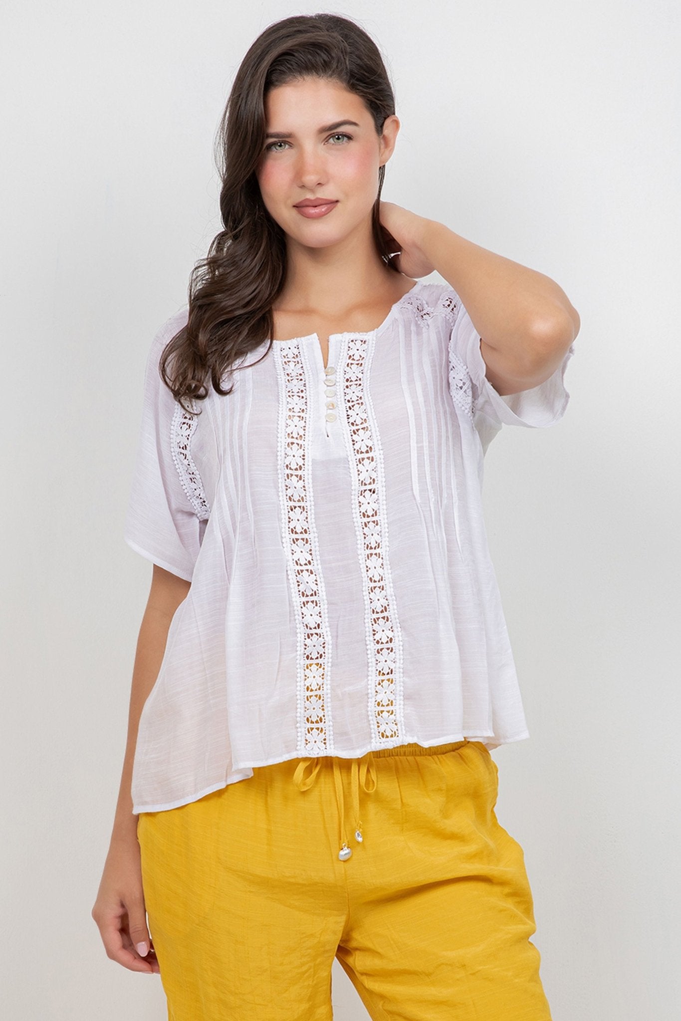 Woman wearing the white Ladies Boho Pleated Lace Panel Blouse, featuring lace panels and short sleeves, with mustard-colored pants.