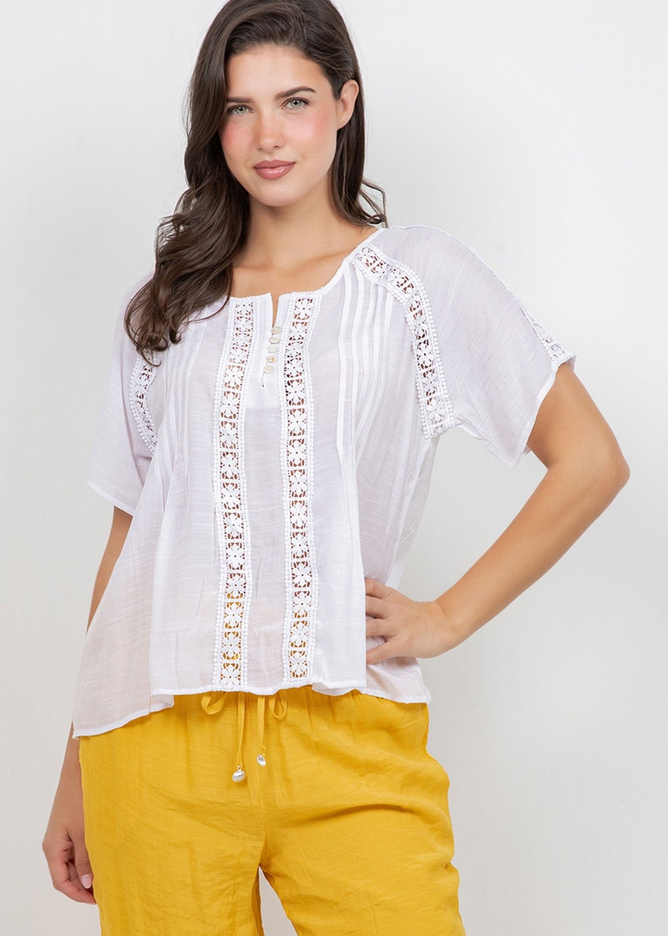 A woman models the white Ladies Boho Pleated Lace Panel Blouse with yellow pants.