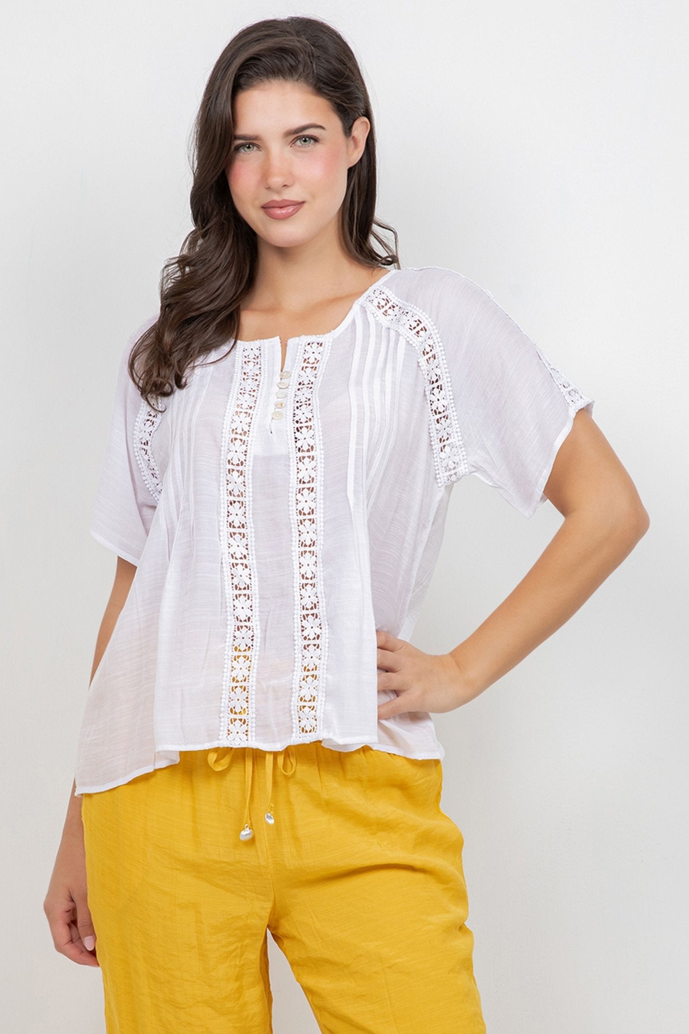 A woman models the white Ladies Boho Pleated Lace Panel Blouse with yellow pants.