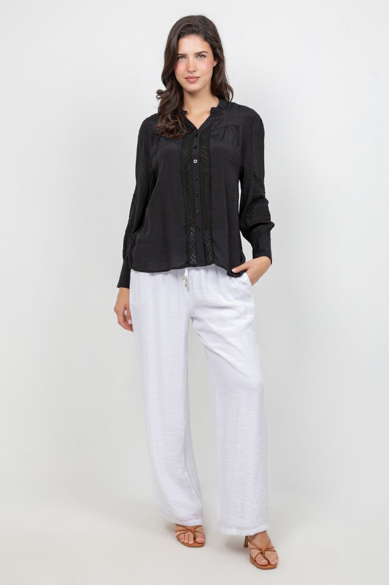 A model wears the black Ladies Boho Lace Panel Pleated Long Sleeve Blouse with white pants in a full studio shot.