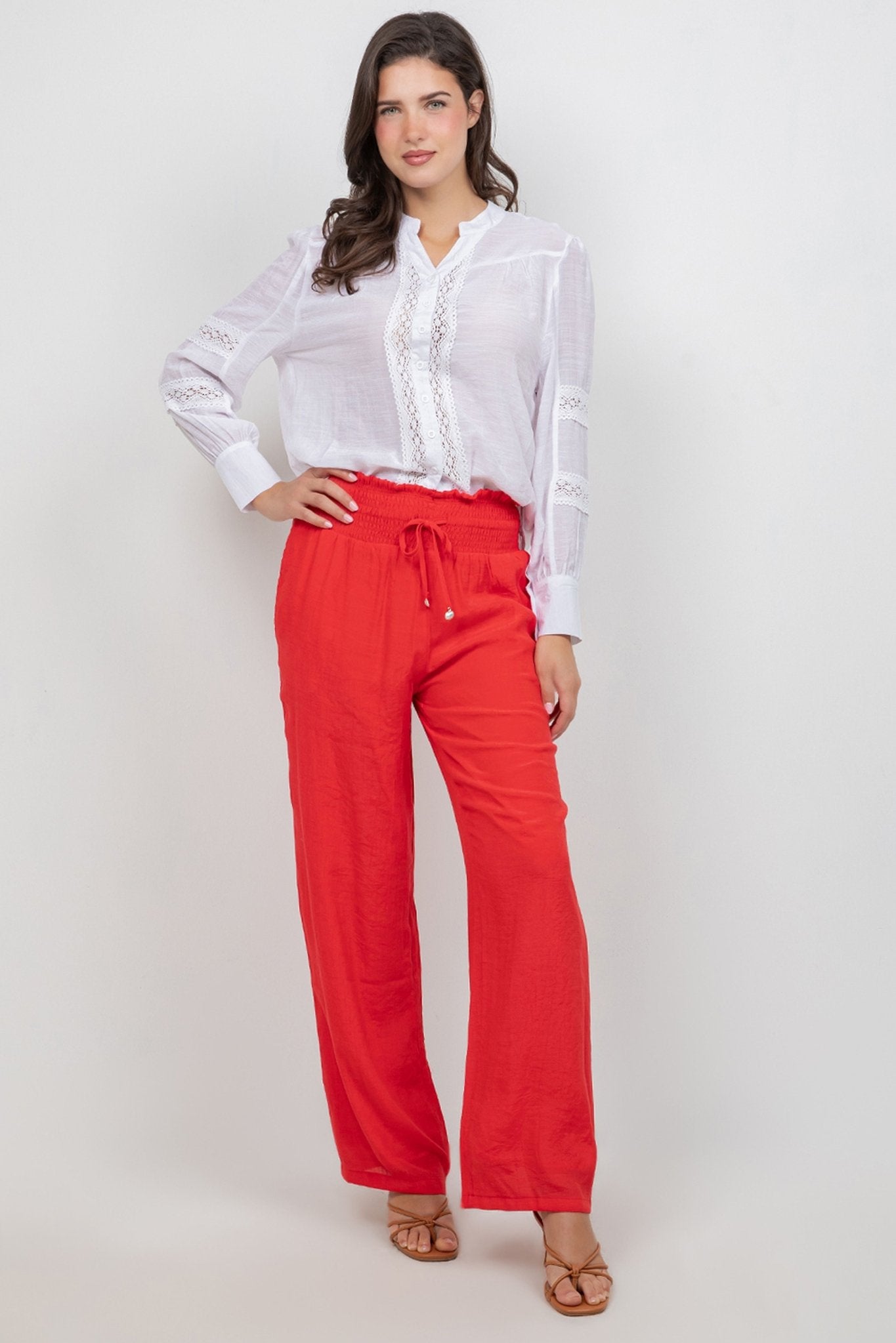 Front-facing view of a model wearing the white Ladies Boho Lace Panel Pleated Long Sleeve Blouse and red pants.